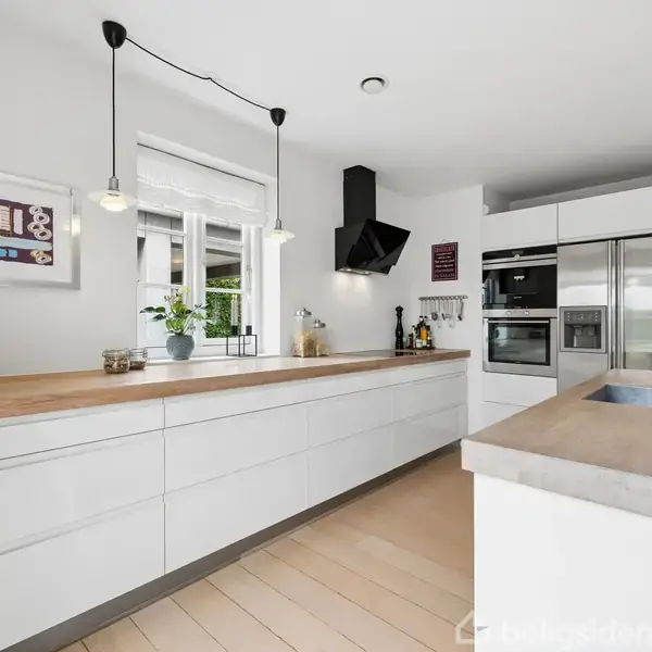 Moderne køkken med hvide skabe har indbygget ovn og køleskab. En blender står på bordpladen, ved siden af en vask og en plante. Væggen bagved har et vindue og et vægmonteret billede.