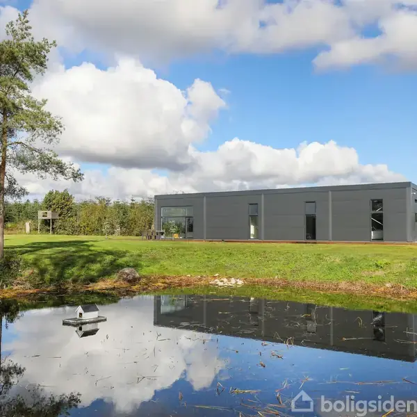 Bygning med grå facade, stående stille, omgivet af græs og et træ. I forgrunden reflekterer en lille dam himmelens skyer og bygningen.