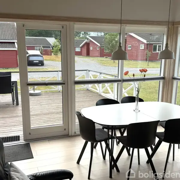Et rundt spisebord med sorte stole står i en lys stue. Gennem store vinduer ses en træterrasse med udendørs møbler og udsigt til røde huse i baggrunden.