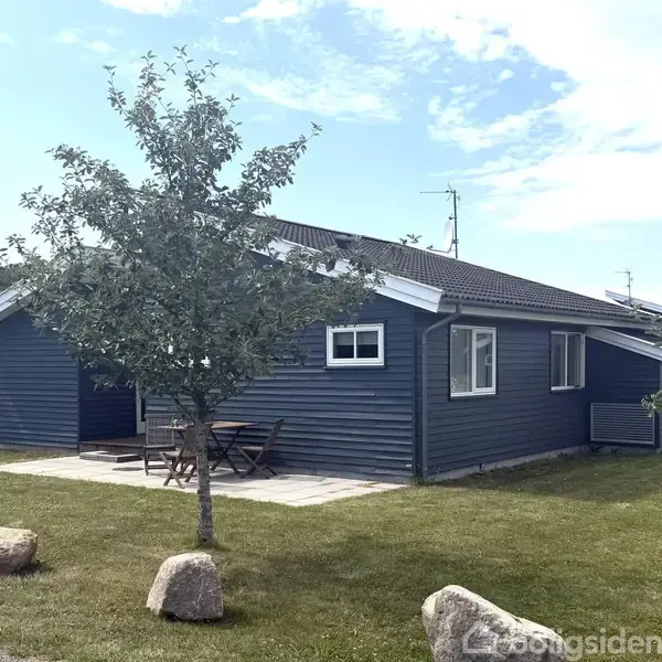 Et blåmalet hus står roligt på en græsplæne med små træer og store sten. Et sæt havemøbler er placeret på en flisebelagt terrasse.