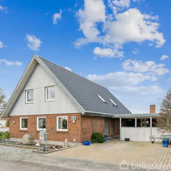 Et hus med spidst tag står på en stille vej. Det har to etager, udvendige murstensvægge og flere vinduer. Omkringliggende træer og en flisebelagt indkørsel skaber en hyggelig atmosfære.