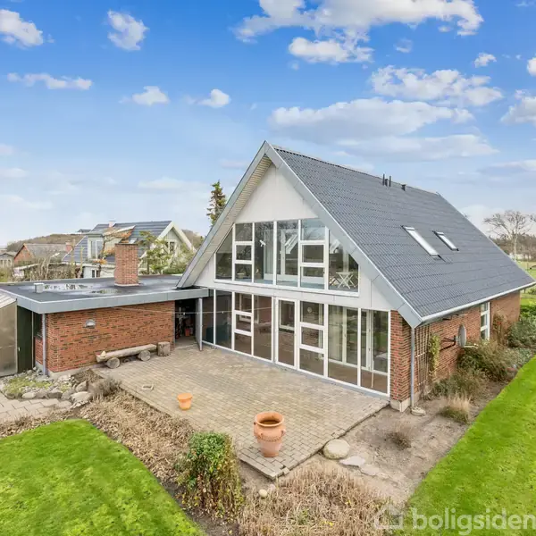 Et rødt murstenshus med en stor terrasse og store vinduer. Det står i en have med græsplæne. Himlen er blå med få skyer, og huset har et skifertag.