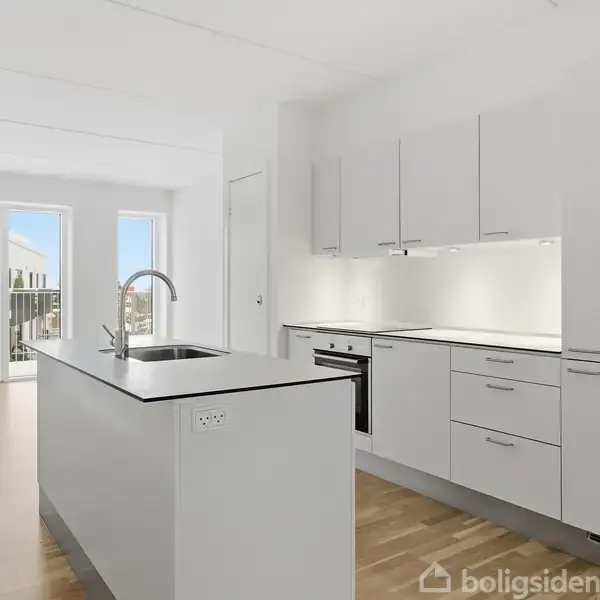Moderne køkken med hvide skabe og en central ø med vask; trægulv løber gennem rummet. Store vinduer i baggrunden giver naturligt lys indad og udsigt til en altan.
