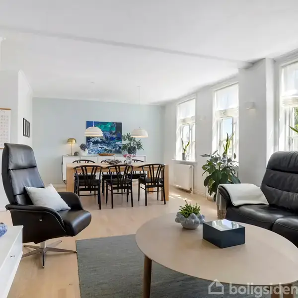 Stole og bord står i et lyst stueområde med store vinduer. Bagved ses et spisebord og stole. Grønne planter tilføjer liv til rummet. En fladskærm hænger på væggen.