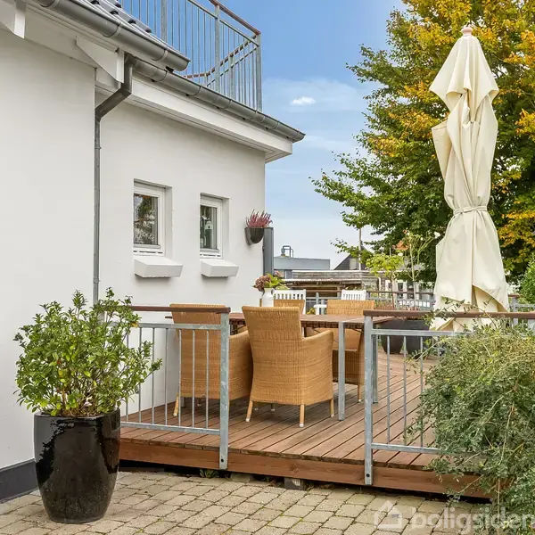 En terrasse med et bord og flettede stole står ved et hvidt hus. En stor parasol er lukket. Grønne buske og et træ med efterårsfarver omkranser området.