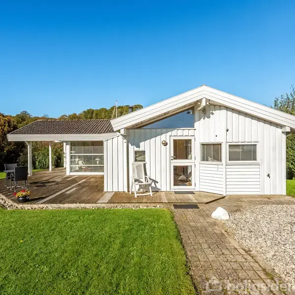 Huset er hvidt med en træterrasse og to hvilestole foran. Solrigt vejr omgiver huset, mens det ligger i en grøn have med en klar blå himmel.