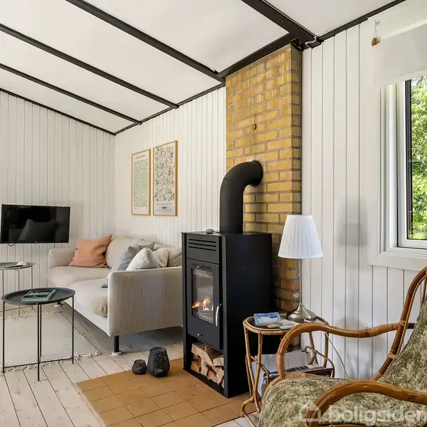 Pejs varmer stue med hvidmalede trævægge, sofa, tv på væggen, og stort vindue mod have. En kurvestol står ved siden af pejsen, der også har et lille indbygget brændrum.