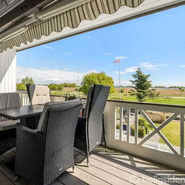 En terrasse med et udendørs bord og stole står under en stribet markise. I baggrunden ses en grøn have med træer og marker under en blå himmel.