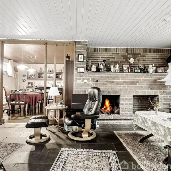 Pejsen brænder i stuen med en murstensvæg som baggrund. Lænestole og sofabord står på tæpper. Køkkenet ses gennem en åben træskillevæg. Lamper hænger fra loftet.