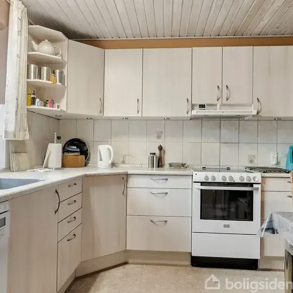 Et køkken med hvide skabe og et gaskomfur står i hjørnet. Ved siden af er der en opvaskemaskine til venstre og en kaffemaskine til højre. Bord med stol i forgrunden.