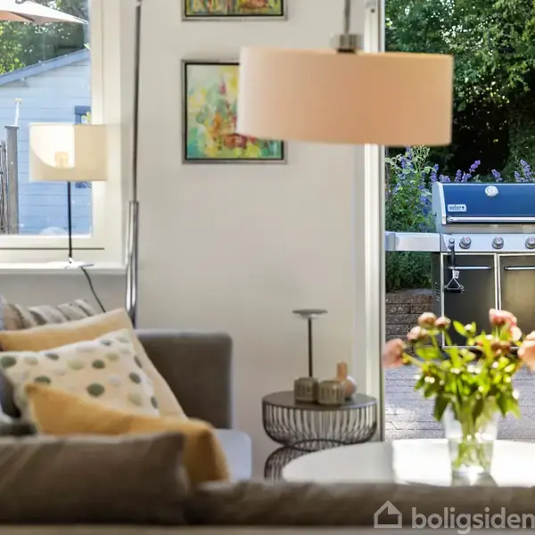 Sofa med puder står i en stue, belyst af en gulvlampe. I baggrunden er en have med en grill og blomster. Vase med blomster ses på et sofabord.