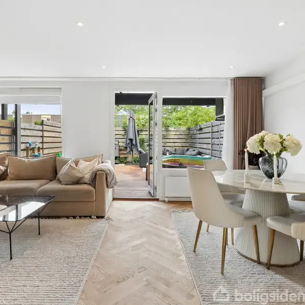 En moderne stue med en beige sofa og et sofabord til venstre, åbne terrassedøre i midten, og et rundt spisebord med blomster til højre. Udenfor ses en terrasse.