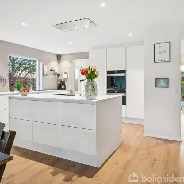 Køkkenø med hvide skuffer står i et lyst køkken. Blomster i en vase pryder øen. Plantegninger hænger på væggen. Store vinduer giver udsigt til træer udenfor.