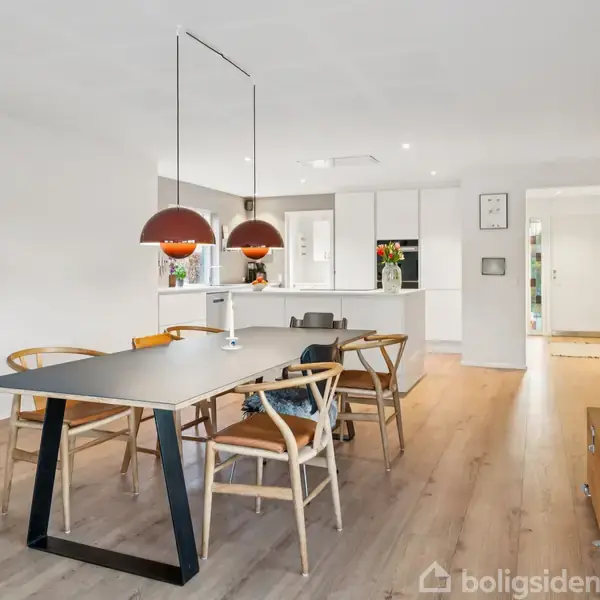 Et spisebord med stole står i et lyst køkkenalrum. To røde lamper hænger over bordet. Køkkenet har hvide skabe, en ø og blomster i en vase.