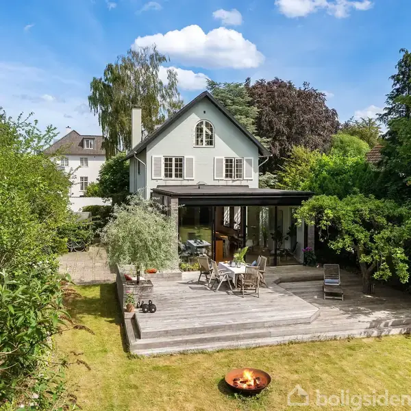 Et lysegrønt hus med skråt tag står omgivet af frodig vegetation. En stor, træbelagt terrasse foran huset har borde, stole og planter. Græsplænen omkring er velplejet og grøn.
