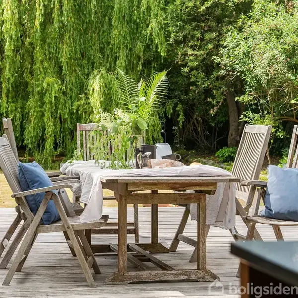 Træbord med høje stole, dækket med dug og blomsterdekoration, står på en terrasse ved siden af en frodig have med træer og buske.