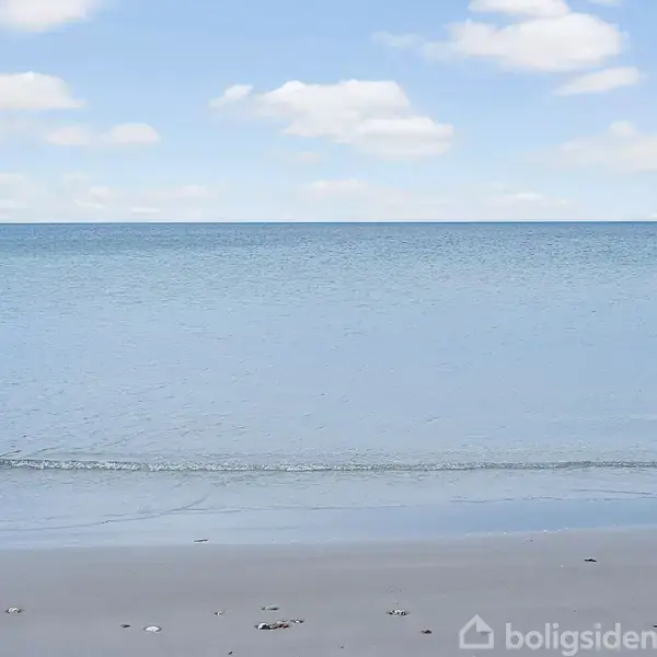 Strand ved vandkant med lette bølger; klar himmel med hvide skyer i baggrunden.