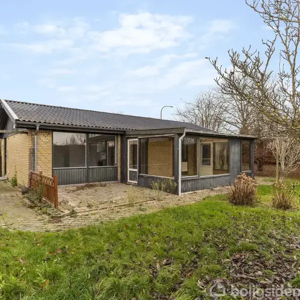 Et enkelt hus med murstensfacade og mørkt tag har en tilstødende terrasse. Det står i en have med græsplæne, træer og buske under en klar himmel. Ingen tekst ses.