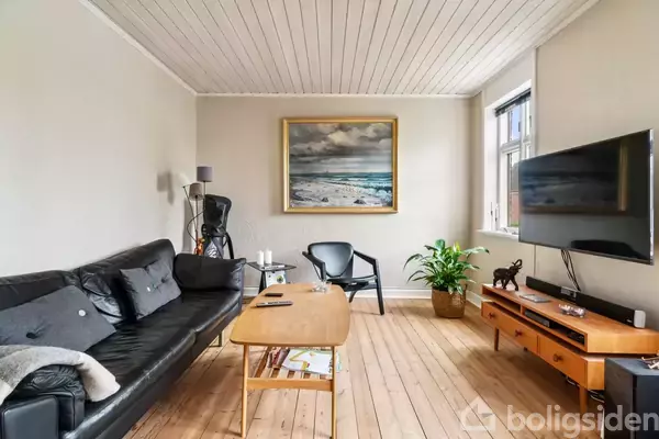 En stue indeholder en sort lædersofa, der står ved et træbord med bøger under, omgivet af en stol, et maleri, en plante og et væghængt TV.