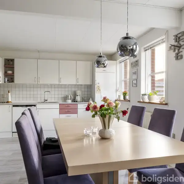 Et spisebord med blomster i en vase står i et lyst køkken. Køkkenet har hvide skabe, to hængelamper, og store vinduer, der slipper sollys ind.