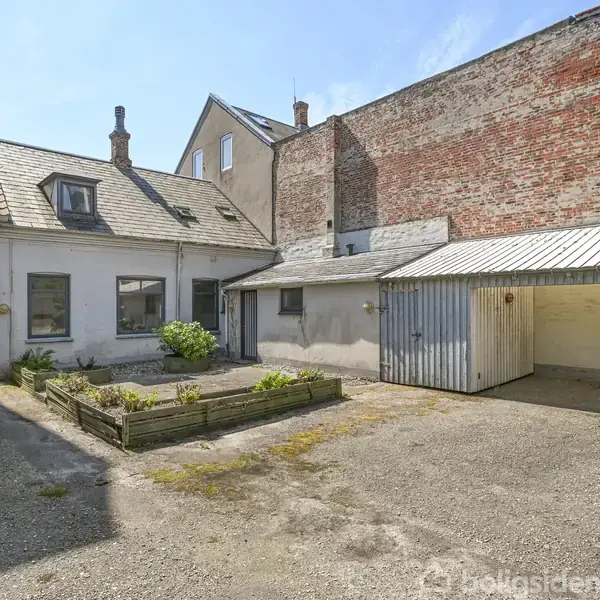 Hus med skråt tag og kviste står i en gård, ved siden af et åbent skur. Gården er omgivet af mure og indeholder et mindre bed med planter.