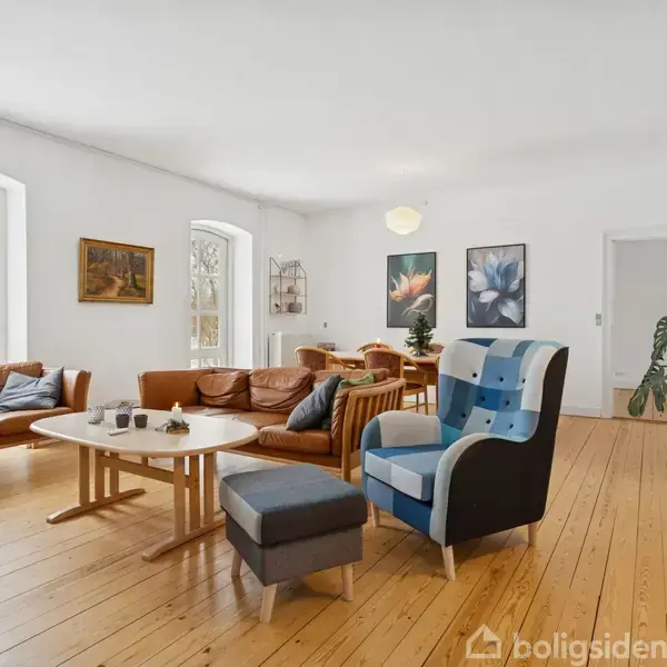 Stue med lyse trægulve og hvide vægge. Sofa, lænestol og sofabord står ved vinduet. Spisebord og stole i baggrunden. Væghængte billeder. Store buede vinduer giver lys.