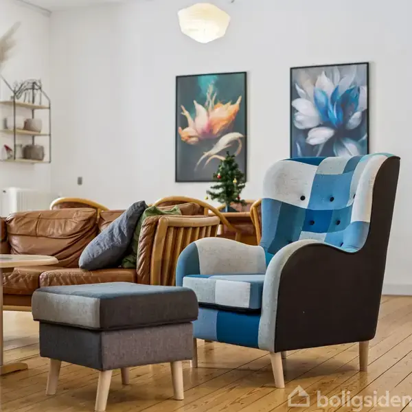 En patchwork-lænestol med fodskammel står på et trægulv i en stue. Bagved ses en brun lædersofa og farverige blomstermalerier på væggen. En lille, dekoreret plante er placeret ved sofaen.