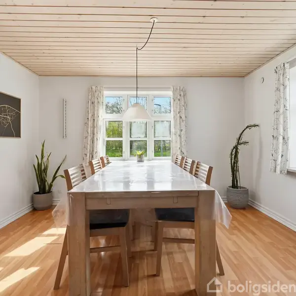 Langt spisebord med seks stole står på trægulv. En loftslampe hænger over bordet. Væggene er hvide med planter og et vægkort. Vinduesudsigt med solskin.
