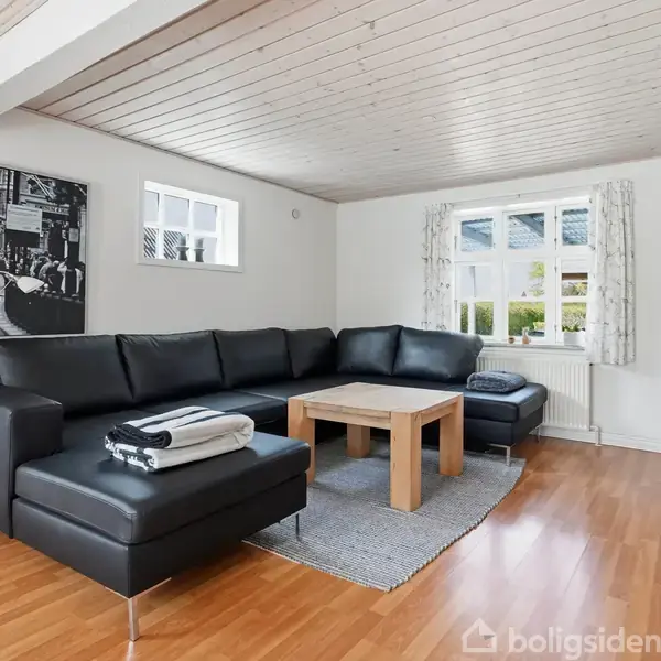 Sort lædersofa står i hjørnet af en lys stue med trægulv og hvidt loft. Et stort sort-hvidt billede hænger på væggen, og der er et sofabord foran sofaen.