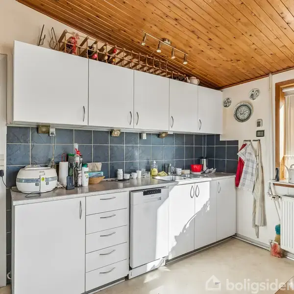 Køkken med hvide skabe og opvaskemaskine under en træbeklædt loft. Væggen har blå fliser, og der er en stor vindue med udsigt til træer. Bordet er fyldt med køkkenudstyr.