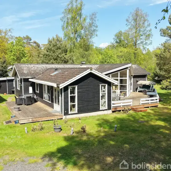 Sort træhus står på en græsplæne omgivet af træer. Terrassen foran huset har udendørsmøbler, mens huset har store vinduer og skråt tag.