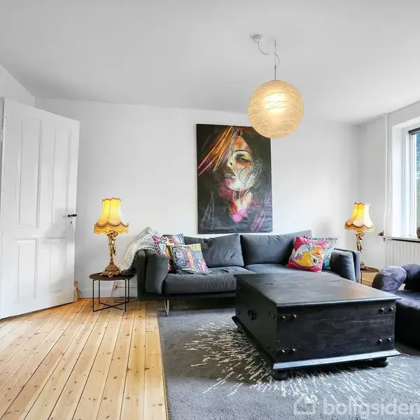 En stue med en grå sofa er dekoreret med farverige puder og en stor kunstplakat på væggen. Et rundt lampehænger i loftet. Store vinduer lukker lys ind.