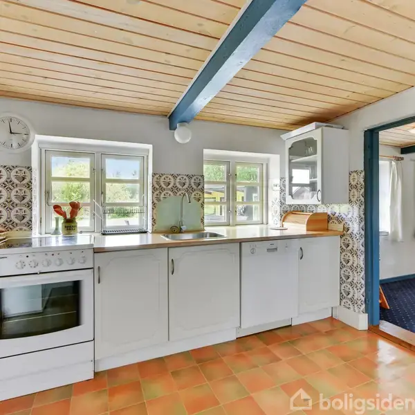 Et køkken med hvide skabe og en komfur, står i et rum med flisebelagt gulv og dekorative fliser på væggene. Tre vinduer tillader lys at strømme ind.