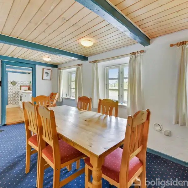 En træspisebord med seks stole står i midten af et rum. Ild i pejsen til venstre. Møblerne er omgivet af hvide vægge og vinduer med hvide gardiner.