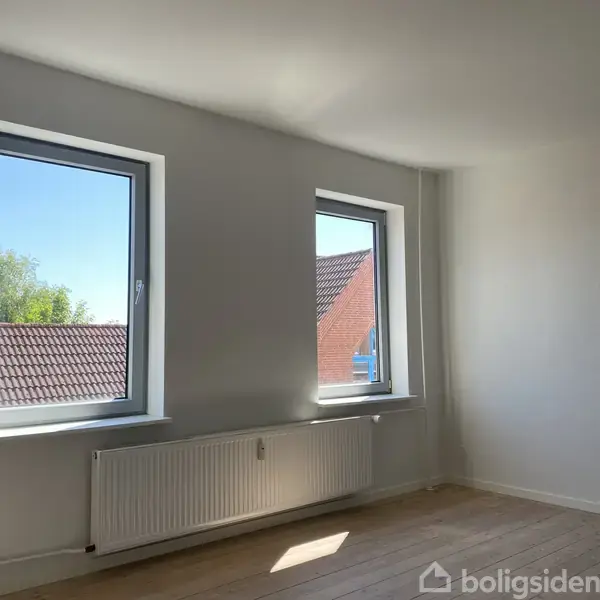 To vinduer lader lys ind i et rum med hvide vægge og trægulv. En radiator er placeret under vinduerne. Udsigten viser et tag og træer udenfor.