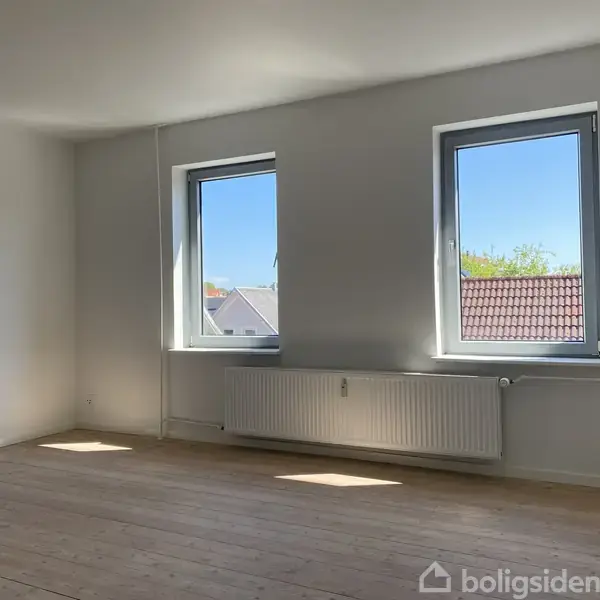 To store vinduer lader sollys strømme ind i et tomt værelse med trægulv og radiator under vinduerne. Udsigten viser tage og blå himmel.