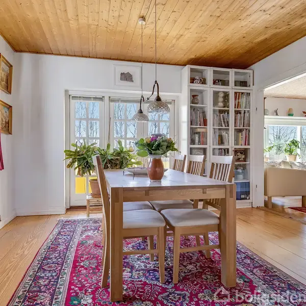 Spisebord med seks stole står på en rød orientalsk tæppe. Bordet har blomstervase. Store vinduer lukker lys ind, og en bogreol står ved siden af en indgang til en stue.