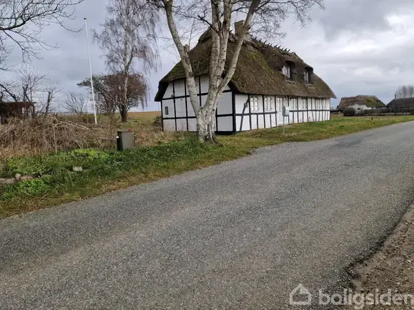 Bindingsværkshus med stråtag står ved vejkanten. Træer omgiver bygningen i landligt område med grusvej og markjord.