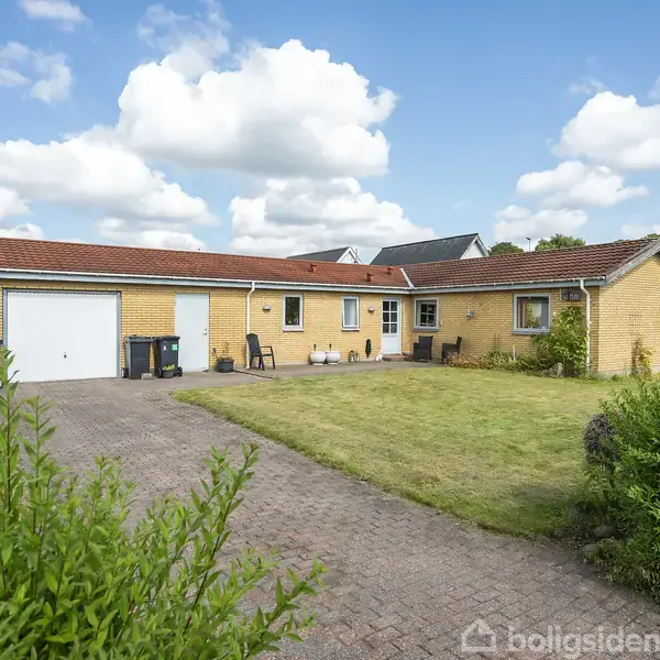 Et gult murstenshus med rødt tegltag står i en velholdt have med græsplæne og flisebelagt indkørsel. Der er nogle havemøbler foran huset under en blå himmel med hvide skyer.