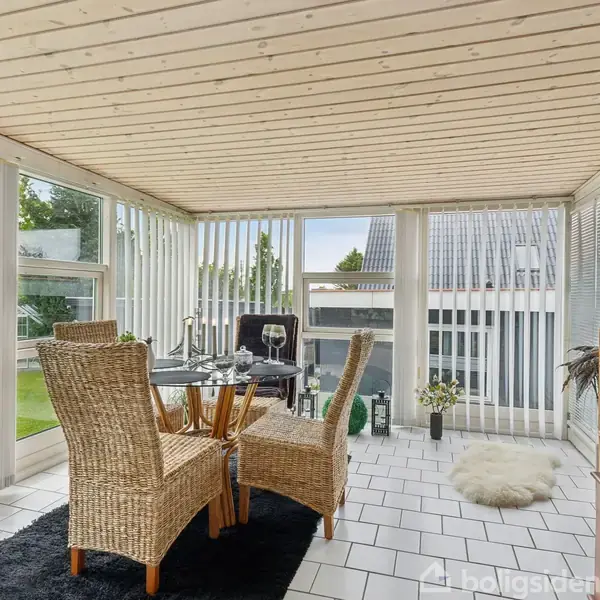 Fletstole står rundt om et spisebord med glasplade, på en indendørs terrasse med lyse trævægge og stort vinduesparti. Der er planter og dekorationer i rummet.