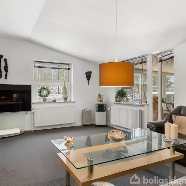 En moderne stue med sort lædersofa og glas-sofabord. En stor orange pendellampe hænger over bordet. Væggene er hvide, og der er en fladskærm på væggen samt vinduer med planter.