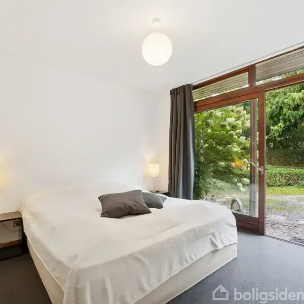 Seng i et soveværelse med gråt tæppe, flankeret af to små lamper. Store vinduer med udsigt til en have med buske og træer.