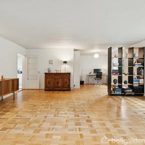 En rummelig stue med trægulv, en skænk til venstre og en reol med bøger til højre. Et skrivebord med stol ses i baggrunden nær et stort vindue.