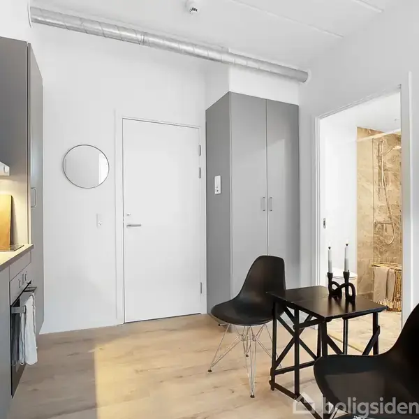 Et moderne køkken med grå skabe og stålvask. To sorte stole står ved et lille bord i centrum. Et spejl hænger på væggen, og en dør fører til et lysere rum.