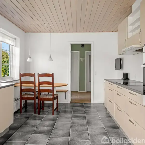 Køkken med beige skabe og moderne apparater, herunder ovn og kogeplader. To træstole og et bord står ved et vindue. Hvidmalede vægge og mørkt flisegulv skaber en lys atmosfære.
