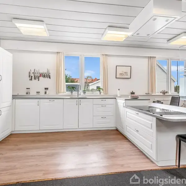 Et rummeligt køkken med hvide skabe og en indbygget ovn. En stor køkkenø med kogeplader og sorte stole omkring. Lysindfald fra flere vinduer i baggrunden.