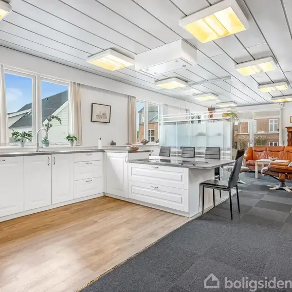 Kontorområde med hvide skabe, sort stol ved computerbordet, og brune lædersofaer danner en siddegruppe. Store vinduer giver naturligt lys; loftslamper oplyser rummet.