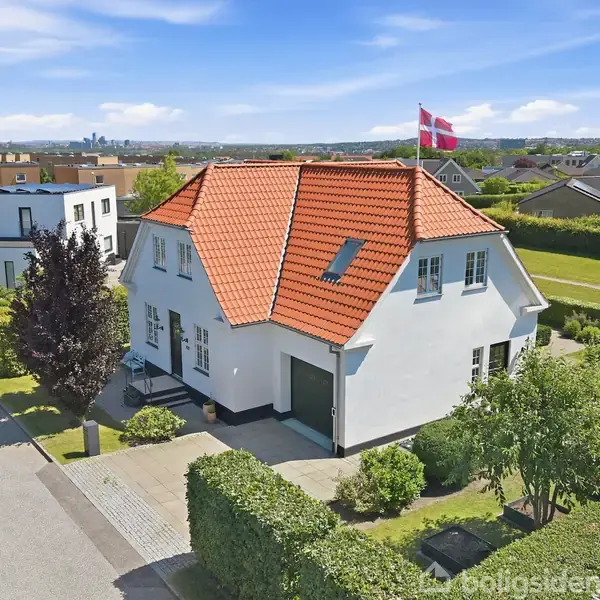 Hus med rødt tegltag og hvide vægge, der står i en velplejet have. En dansk flag vajer på taget. I baggrunden ses et boligkvarter under en blå himmel.