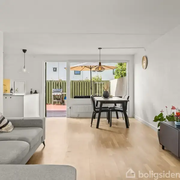 Stue med grå sofa, der står stille i et lyst rum. Et spisebord i mørkt træ står tæt på en åben dør, der fører til en terrasse. TV og planter på skænk.