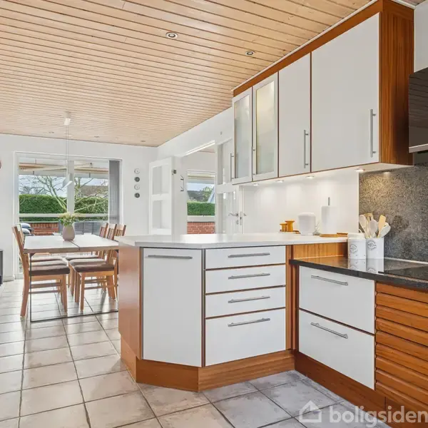 Køkken med hvide og træfarvede skabe, køkkenø med skuffer, og keramiske kogeflade. Spisebord med stole i baggrunden. Stort vindue med udsigt til have. Flisebelagt gulv og træloft.